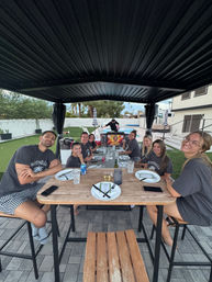 Smiling group of friends seated around a wooden outdoor dining table under a black pergola next to a backyard pool, ready for a casual meal with plates, drinks and a barbecue in the background.