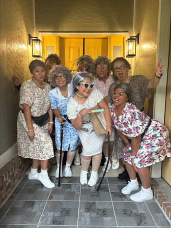 Group of eight adults in playful 'old lady' costumes posing on a front porch in front of a bright yellow door — floral dresses, gray wigs, reading glasses, pearl necklaces, canes and white sneakers, goofy expressions and heart-shaped sunglasses.