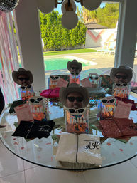 Poolside bachelorette party gift table with cowboy hats, sunglasses, iridescent gift bags and pajamas beside a backyard swimming pool