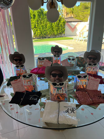 Poolside bachelorette party gift table with cowboy hats, sunglasses, iridescent gift bags and pajamas beside a backyard swimming pool