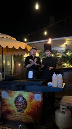 Nighttime outdoor patio hibachi show with two chefs at a mobile griddle under string lights; one wears a tall hat topped by a flame, squeeze bottles and propane tank nearby.
