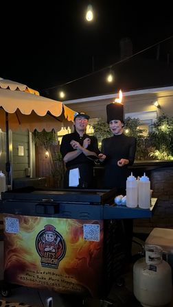 Nighttime outdoor patio hibachi show with two chefs at a mobile griddle under string lights; one wears a tall hat topped by a flame, squeeze bottles and propane tank nearby.