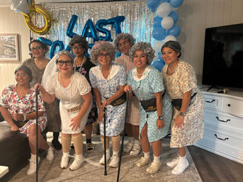 Eight women in playful elderly-themed costumes with gray wigs, floral dresses, pearls, fanny packs and canes posing in a living room in front of blue-and-white balloons, silver fringe backdrop and a gold ring balloon for a bridal shower celebration.