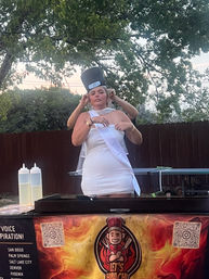 Person in a white dress and sash having a tall chef hat adjusted while standing behind an outdoor hibachi grill cart with flame logo, trees and wooden fence at sunset