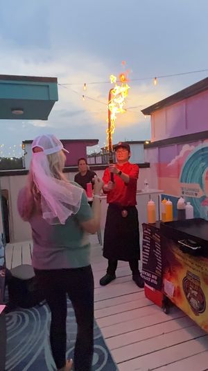 Rooftop deck at dusk with string lights; a cook holding a tall flaming torch near a grill while a guest wearing a white veil and baseball cap watches, colorful mural and condiment bottles nearby.