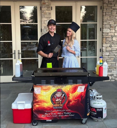 Smiling hibachi chef in black uniform and woman in a striped dress with a tall chef hat posing behind a portable hibachi griddle cart on a stone patio, with condiment bottles, cooler and propane tank visible.