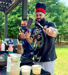 Two cooks at a sunny park barbecue behind a flat-top griddle, one with a tall chef hat and beard holding a spatula and fork; stacked plastic prep containers, squeeze bottles and lush green trees in the background.