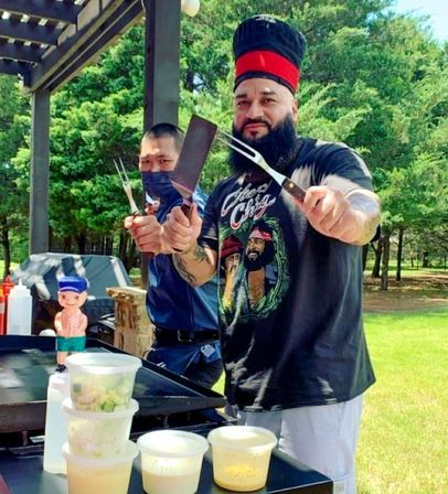 Two cooks at a sunny park barbecue behind a flat-top griddle, one with a tall chef hat and beard holding a spatula and fork; stacked plastic prep containers, squeeze bottles and lush green trees in the background.