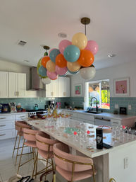 Sunlit modern white kitchen decorated for a celebration with a pastel balloon cluster suspended over a long island, pink velvet bar stools, glasses and snacks on the countertop, teal subway-tile backsplash and a window looking to the backyard.