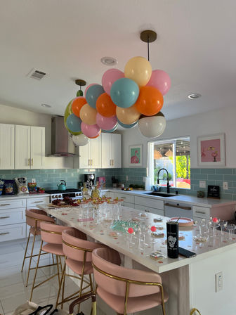 Sunlit modern white kitchen decorated for a celebration with a pastel balloon cluster suspended over a long island, pink velvet bar stools, glasses and snacks on the countertop, teal subway-tile backsplash and a window looking to the backyard.
