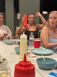 Casual backyard patio dinner with friends around a table of blue plates and bowls; woman playfully raises a red squeeze bottle, red condiment bottle in foreground.