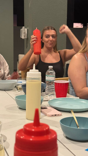 Casual backyard patio dinner with friends around a table of blue plates and bowls; woman playfully raises a red squeeze bottle, red condiment bottle in foreground.