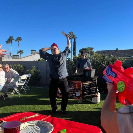 Sunny backyard party with palm trees; a cook in a red cap theatrically raises a spatula at a portable grill while laughing guests aim a red water gun.