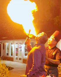 Dramatic fire-breathing: a man exhales a massive flame while a chef in a tall hat assists on an outdoor restaurant patio at night.