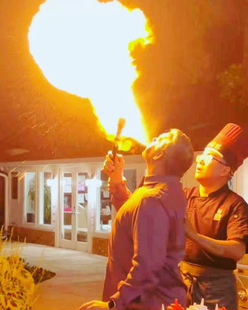 Dramatic fire-breathing: a man exhales a massive flame while a chef in a tall hat assists on an outdoor restaurant patio at night.