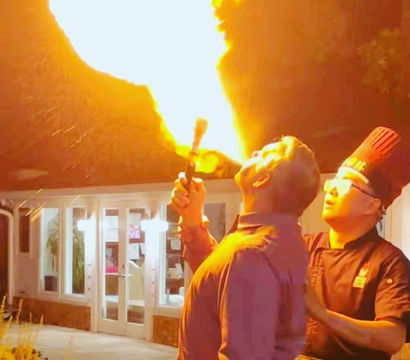 Man performing dramatic fire-breathing with a giant flame on an outdoor restaurant patio at night while a chef in a tall hat watches