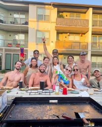 Smiling group of friends at a summer poolside BBQ in front of modern apartment balconies — some shirtless, holding colorful water guns and drinks around a griddle with plates and cups.