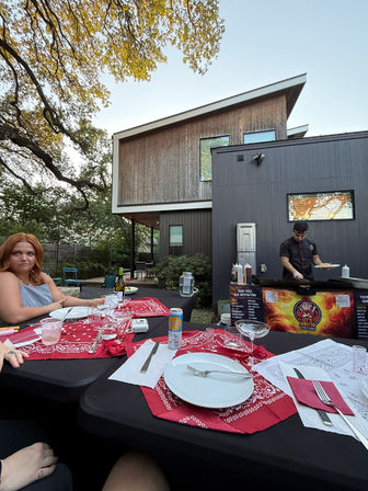 Modern backyard patio dinner at dusk with a chef cooking at a mobile hibachi grill, table set with white plates and red bandana napkins, guests seated under oak tree branches by a contemporary home.