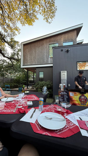 Modern backyard patio dinner at dusk with a chef cooking at a mobile hibachi grill, table set with white plates and red bandana napkins, guests seated under oak tree branches by a contemporary home.