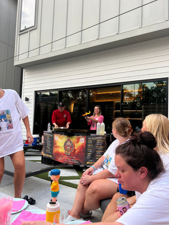 Friends gathered on a modern residential patio watching a chef flambé on a mobile griddle during an outdoor cooking demo, with pink napkins, drinks, and a toy-topped condiment bottle.