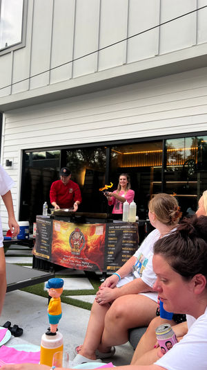 Friends gathered on a modern residential patio watching a chef flambé on a mobile griddle during an outdoor cooking demo, with pink napkins, drinks, and a toy-topped condiment bottle.