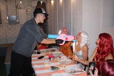 Indoor dinner celebration with friends in colorful wigs, sparklers, and a pink-and-blue toy water gun playfully aimed across a striped tablecloth.