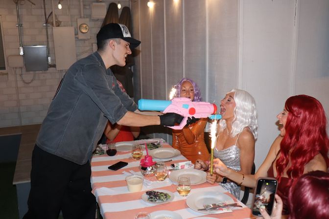 Indoor dinner celebration with friends in colorful wigs, sparklers, and a pink-and-blue toy water gun playfully aimed across a striped tablecloth.