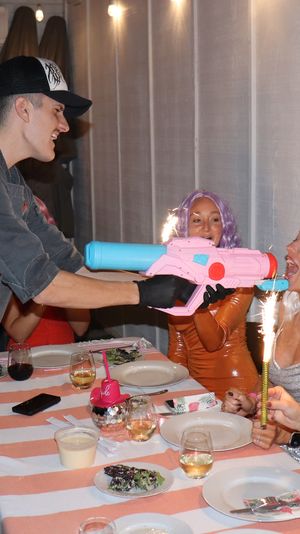 Indoor dinner celebration with friends in colorful wigs, sparklers, and a pink-and-blue toy water gun playfully aimed across a striped tablecloth.