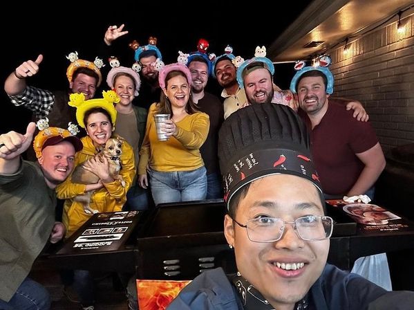 Teppanyaki chef selfie with a smiling group of friends wearing colorful character headbands, one holding a small dog and others raising drinks on a nighttime restaurant patio.