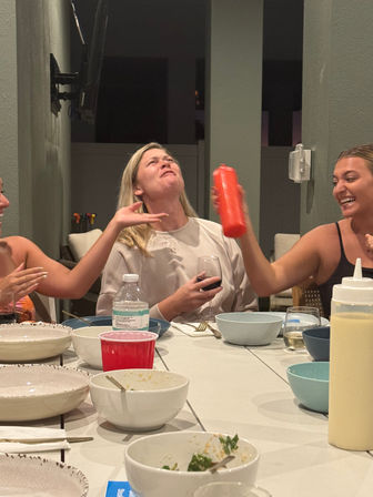 Group of friends at an outdoor patio dinner, one woman playfully squirting a red sauce bottle toward a laughing friend holding a glass of wine, bowls and plates on the table at night.