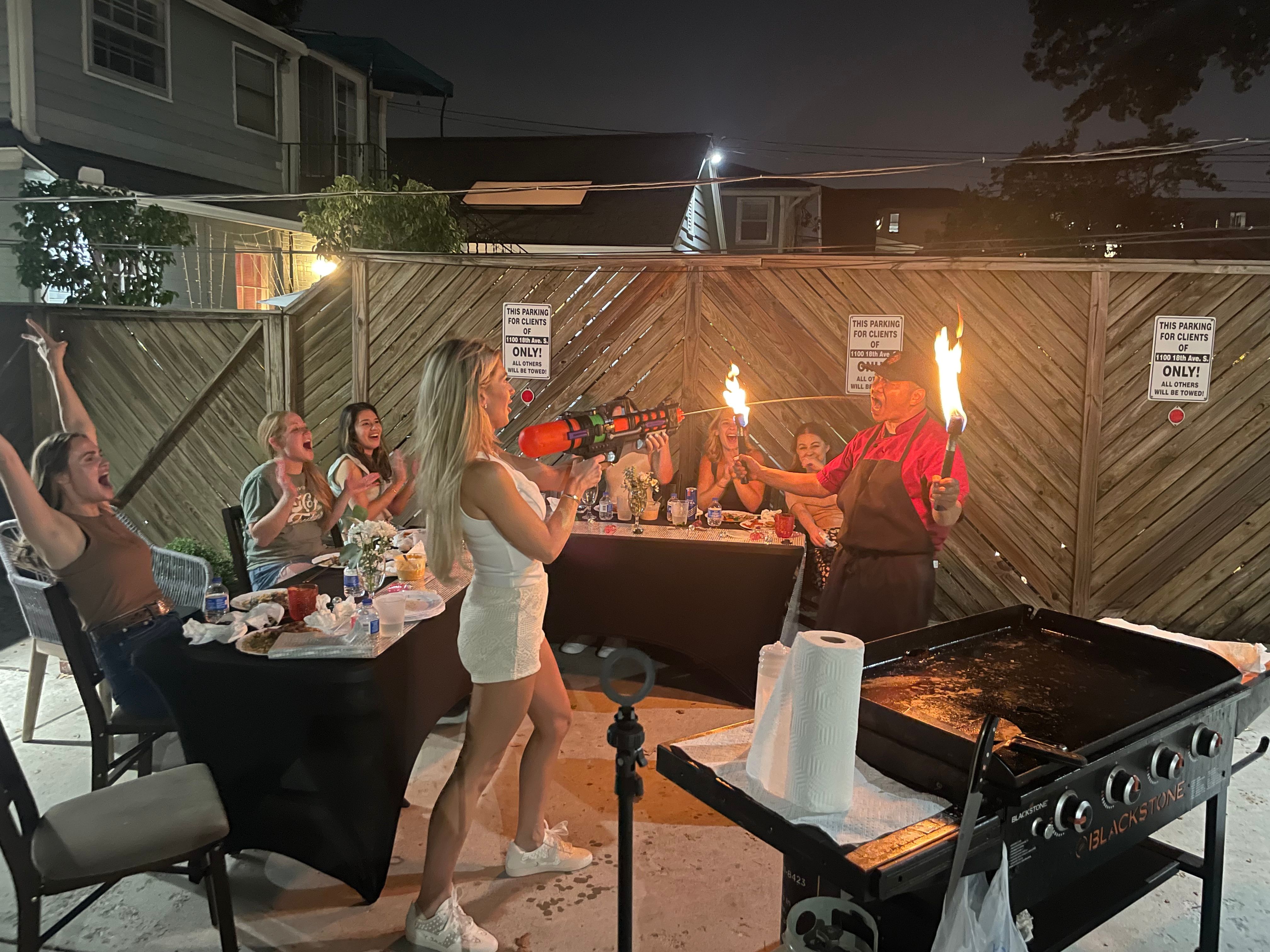 Nighttime backyard patio party with friends cheering as a woman playfully aims a toy blaster at a cook holding two flaming torches beside a large outdoor griddle.