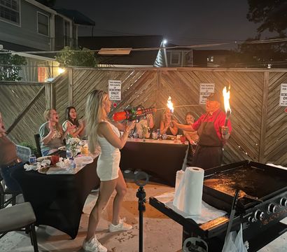 Nighttime backyard patio party with friends cheering as a woman playfully aims a toy blaster at a cook holding two flaming torches beside a large outdoor griddle.
