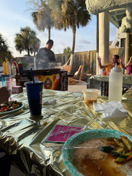 Coastal outdoor dining under palm trees: chef at a smoky grill, gold-covered picnic tables with paper plates, plastic cups and condiments during a relaxed sunset meal.