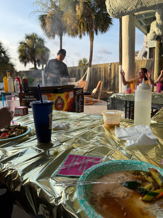 Coastal outdoor dining under palm trees: chef at a smoky grill, gold-covered picnic tables with paper plates, plastic cups and condiments during a relaxed sunset meal.