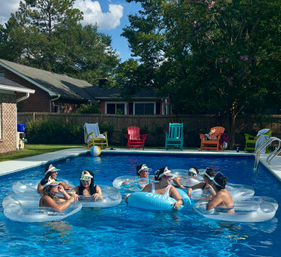 Friends lounging on clear inflatable rings and a blue "Bride" float in a suburban backyard pool on a sunny summer day, with colorful Adirondack chairs, a beach ball, house and trees in the background.