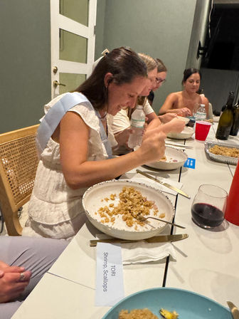 Group of women eating at an indoor dinner party, plates of shrimp and scallop rice on a long table with wine glass, water bottles and a name card labeled "Shrimp, Scallops".