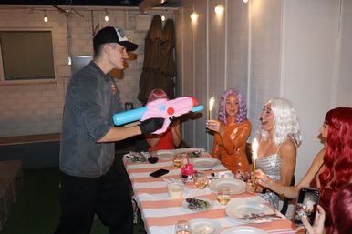Man holding a pink-and-blue toy water gun playfully aims at laughing women in colorful wigs holding cake sparklers around a striped-tablecloth outdoor patio party table.