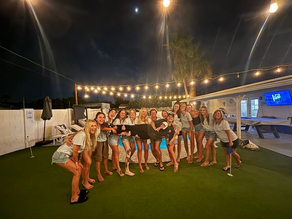 Nighttime backyard pool party: a large group of friends on artificial turf under string lights and a palm tree, laughing as they hold one person horizontally.