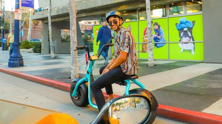 Smiling rider in a helmet and patterned shirt on a teal electric trike scooter on a downtown sidewalk, passing a bright storefront with playful pet murals and a car side‑mirror reflection in the foreground