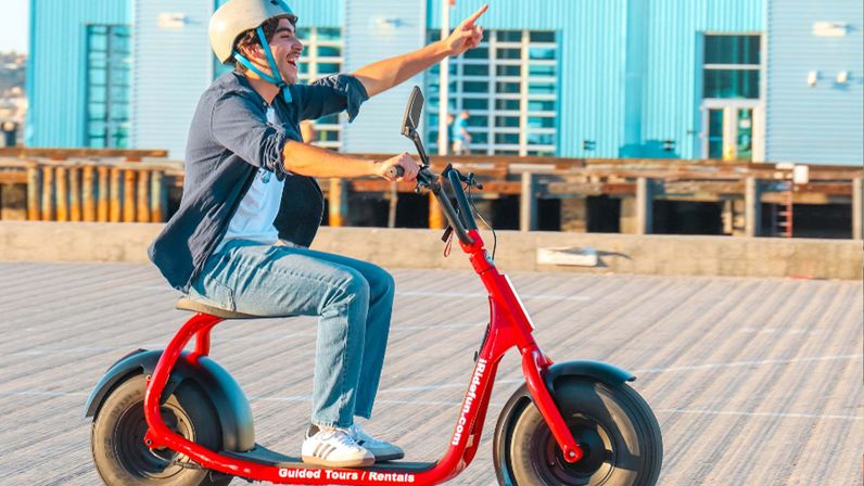 Person wearing a helmet rides a red electric scooter with fat tires along a sunny waterfront boardwalk by blue pier buildings, smiling and pointing ahead.