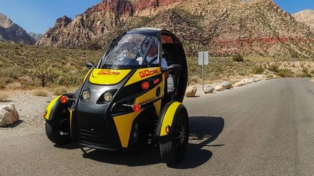 Compact yellow-and-black three-wheeled tour vehicle on a paved desert road with red-rock canyon cliffs and a clear blue sky in the background.