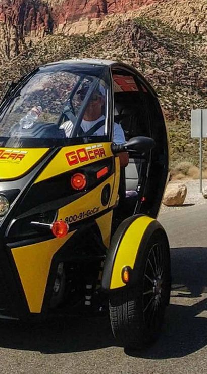 Compact yellow-and-black three-wheeled tour vehicle on a paved desert road with red-rock canyon cliffs and a clear blue sky in the background.