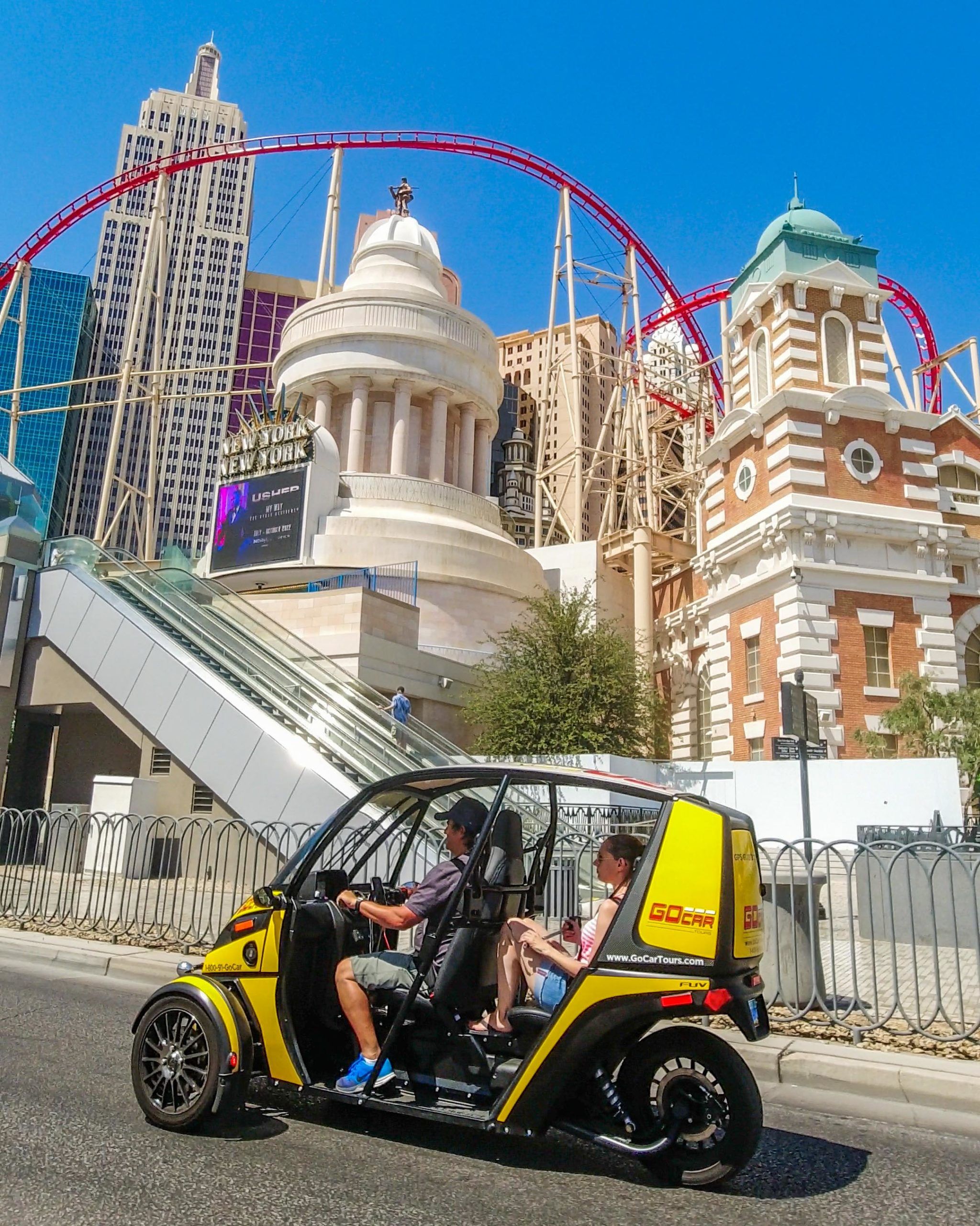 Couple in a bright yellow open-air tour buggy driving on the Las Vegas Strip past New York–style hotel facades and a red roller coaster against a clear blue sky.