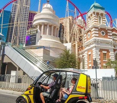 Couple in a bright yellow open-air tour buggy driving on the Las Vegas Strip past New York–style hotel facades and a red roller coaster against a clear blue sky.