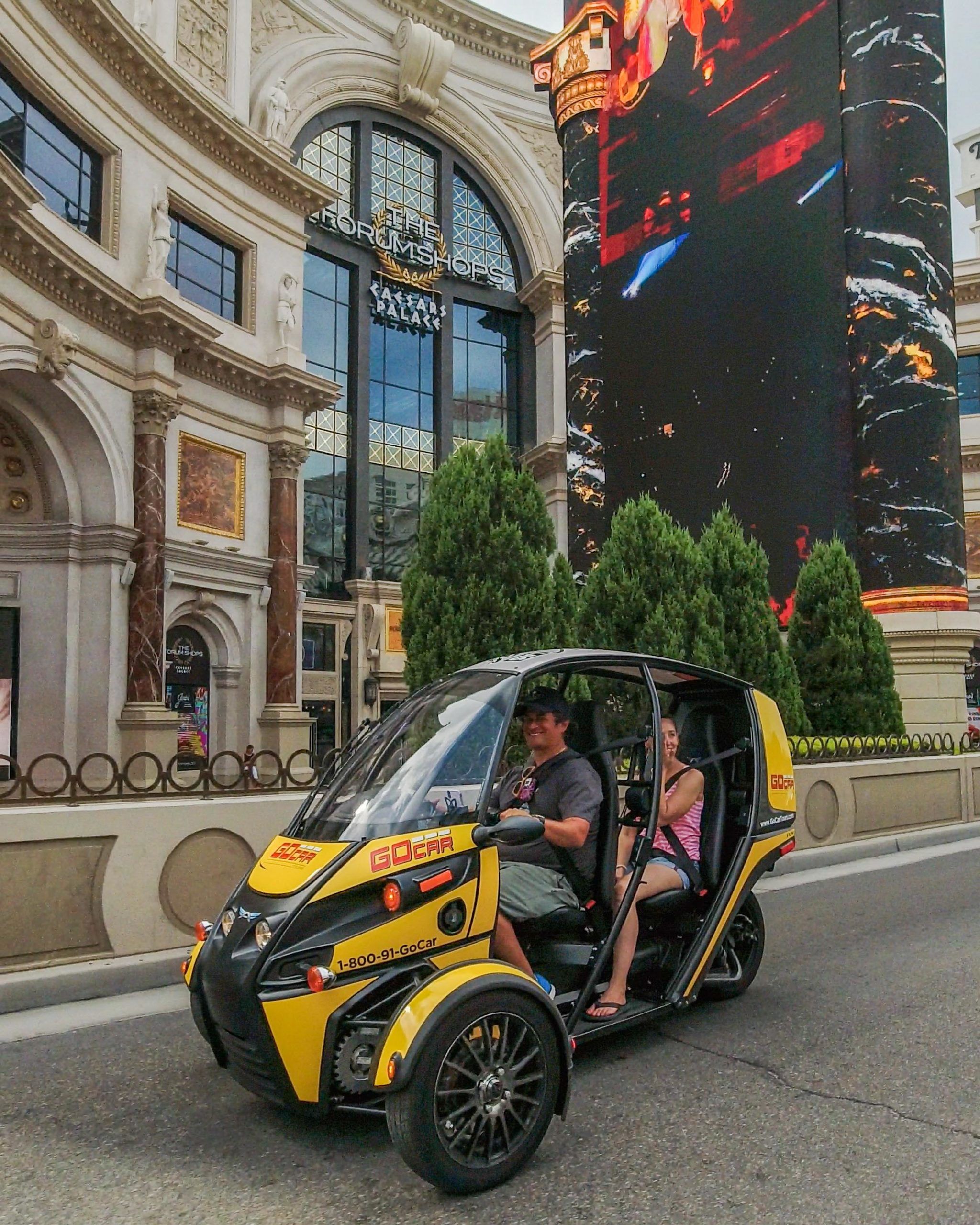 Yellow three-wheeled tour vehicle with two smiling passengers driving past an ornate casino-resort façade and towering digital billboard on the Las Vegas Strip.