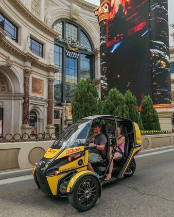 Yellow three-wheeled tour vehicle with two smiling passengers driving past an ornate casino-resort façade and towering digital billboard on the Las Vegas Strip.