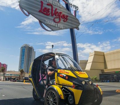 Yellow two-seat rental microcar driving beneath the iconic 'Las Vegas' sign on a sunny day near the Strip, with high-rise hotels and blue sky in the background.
