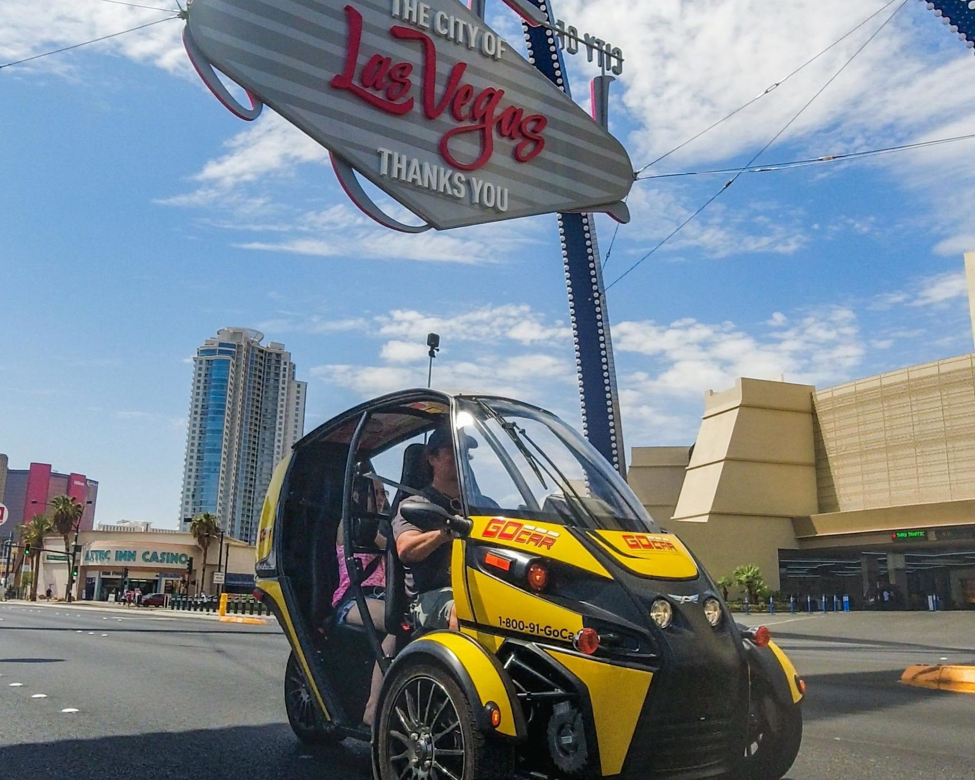 Yellow two-seat rental microcar driving beneath the iconic 'Las Vegas' sign on a sunny day near the Strip, with high-rise hotels and blue sky in the background.