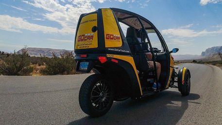Sunny scenic desert road with a yellow three-wheeled tour buggy cruising past shrubs and distant red-rock cliffs under a blue sky