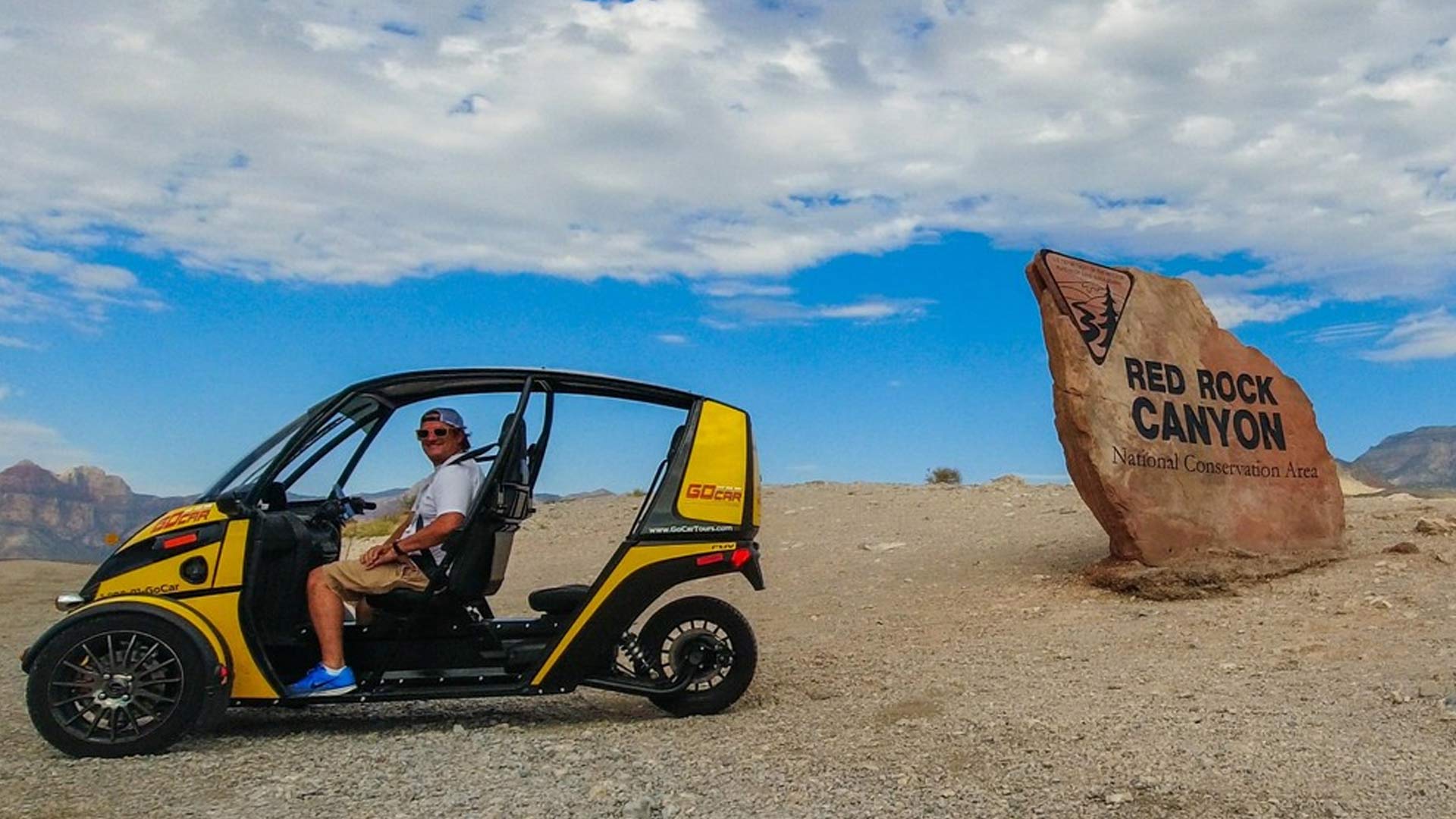 Person in a yellow open-air tour vehicle parked beside a large sandstone sign for Red Rock Canyon National Conservation Area, arid desert landscape and blue cloudy sky